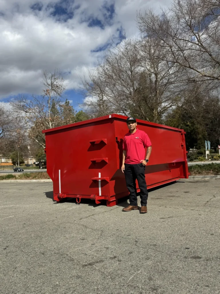 A man in a red polo shirt and cap stands next to a large red dumpster in a parking lot, WeHaul Dumpster Rental